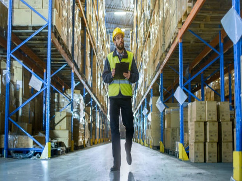 Overseer Wearing Hard Hat with Tablet Computer Counts Merchandise in Warehouse. He Walks Through Rows of Storage Racks with Merchandise.